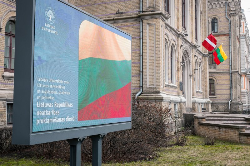 Ekrāns ar sveicienu Lietuvas Republikas neatkarības proklamēšanas dienā un Latvijas un Lietuvas karogi pie Latvijas Universitātes galvenā ēkas. null